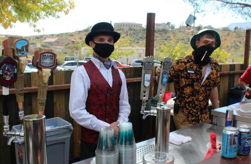 Two bartenders in masks stand behind a bar with beer taps, surrounded by outdoor scenery and colorful cups.