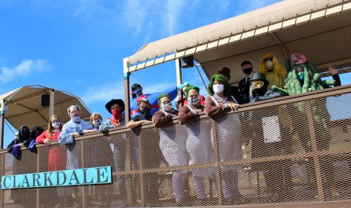 A group of people in costumes and masks poses on a balcony with a sign that reads "Clarkdale."