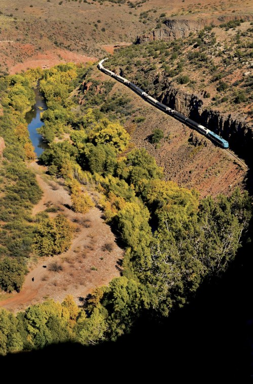 A train winds along a river through a canyon, surrounded by colorful trees and rugged terrain.