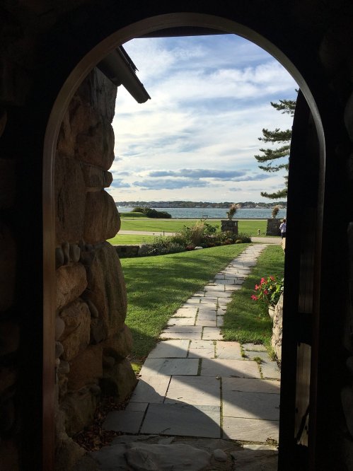 View through an arched stone doorway, showcasing a lush lawn and serene water beyond under a partly cloudy sky.