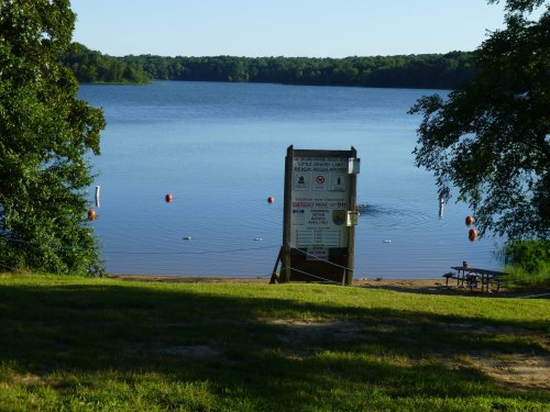 A serene lake view with a signpost near the shore, surrounded by trees and calm water under a clear blue sky.