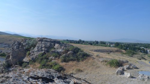 A rocky landscape with sparse vegetation, rolling hills, and distant mountains under a clear blue sky.