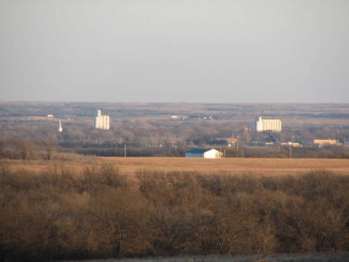 A distant view of a flat landscape with silos and buildings against a hazy sky, surrounded by bare trees.