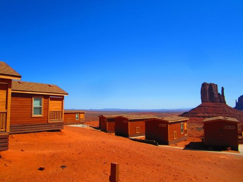 Desert landscape with wooden cabins and a rock formation under a clear blue sky.