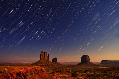 Star trails illuminate the night sky over Monument Valley's iconic rock formations in a serene desert landscape.