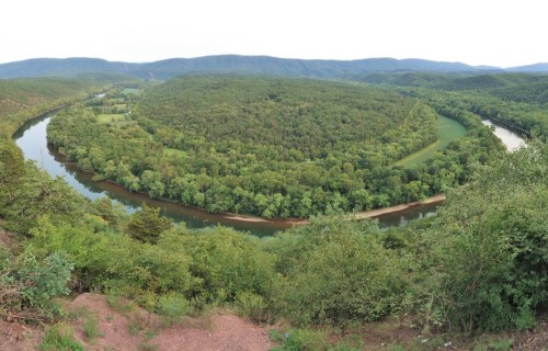 A panoramic view of a winding river surrounded by lush green forests and rolling hills under a cloudy sky.