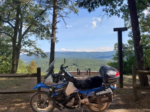 A blue motorcycle parked near a scenic overlook with mountains and trees in the background under a clear sky.