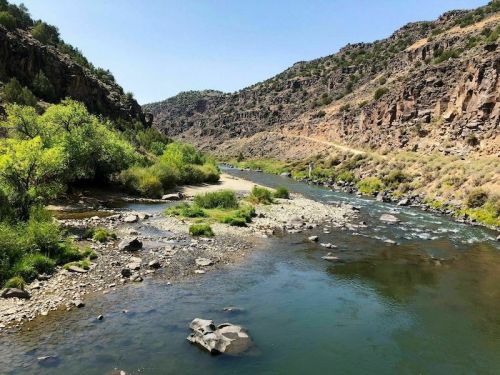 A serene river flows through a rocky canyon, surrounded by lush greenery and rugged hills under a clear blue sky.
