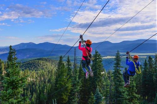 Two people ziplining through a forested area with mountains in the background under a partly cloudy sky.