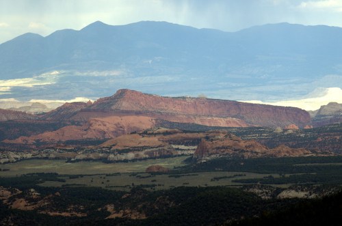 A panoramic view of layered red rock formations and distant mountains under a cloudy sky.