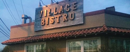 Exterior of Village Bistro, featuring a sign, windows, and surrounding greenery at dusk.