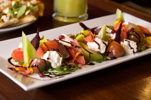 A colorful salad with mixed fruits, cheese, and drizzled balsamic glaze, garnished with edible flowers on a white plate.