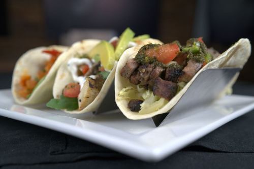 Three tacos on a white plate, filled with grilled meat, fresh vegetables, and garnished with herbs.