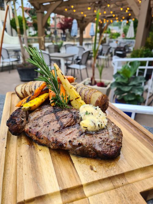 A grilled steak on a wooden platter, topped with herb butter and accompanied by roasted vegetables. Outdoor dining setting in background.
