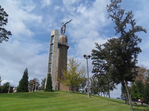 A tall monument with a statue on top, surrounded by trees and a grassy area under a partly cloudy sky.
