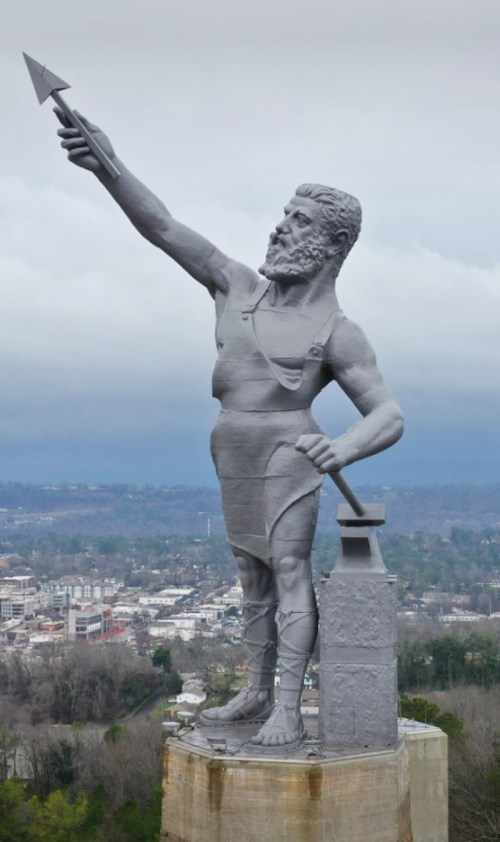 A large gray statue of a muscular figure holding a spear, overlooking a cityscape with cloudy skies.