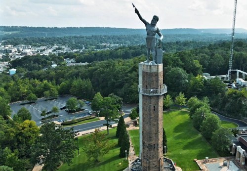 Aerial view of a tall statue atop a brick tower, surrounded by trees and a cityscape in the background.
