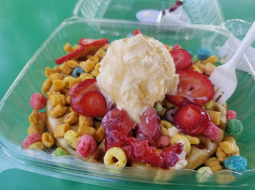 A dessert bowl with a waffle topped with ice cream, strawberries, and colorful cereal pieces.