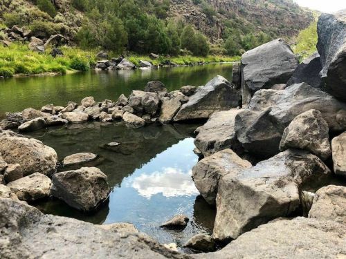 A serene river scene with smooth rocks and clear water reflecting the sky and surrounding greenery.