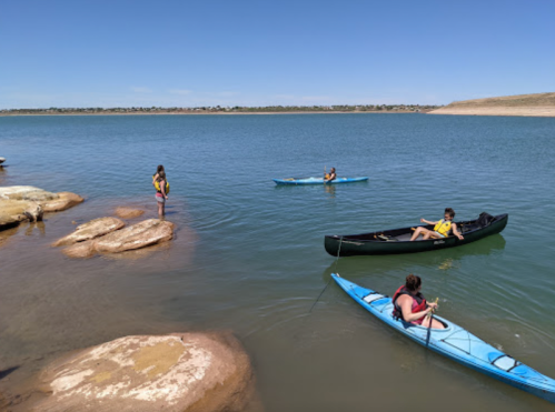 Two kayakers on a calm lake, with a person standing on rocks nearby and another person wading in the water.