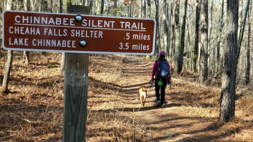 A hiker with a dog walks along the Chinnabee Silent Trail, with a sign indicating distances to Cheaha Falls Shelter and Lake Chinnabee.