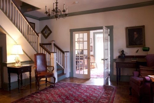 A cozy foyer with a staircase, chandelier, and vintage furniture, leading to a dining area through double doors.