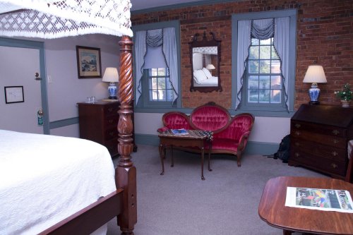 Cozy room with a four-poster bed, vintage furniture, and large windows, featuring a red loveseat and wooden dresser.