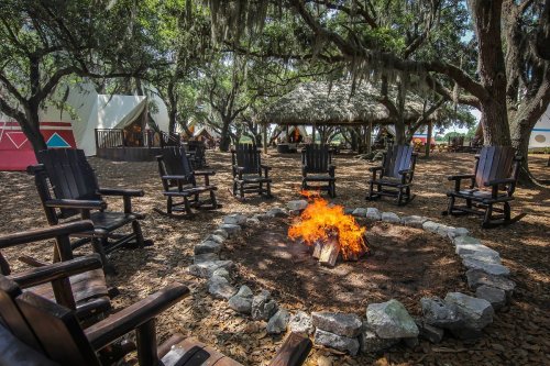 A cozy fire pit surrounded by wooden chairs under trees, with a thatched-roof structure in the background.