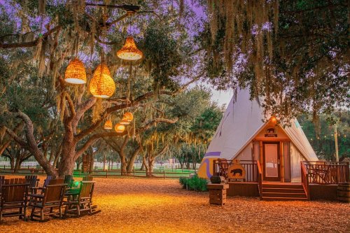 A cozy teepee surrounded by trees, with hanging lights and a rustic seating area on a warm, inviting evening.