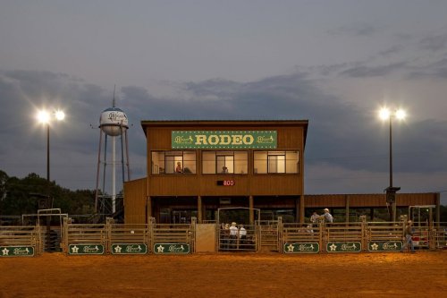 A rodeo arena at dusk, featuring a building with "RODEO" signage and a water tower in the background.