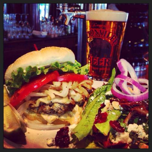 A juicy burger with lettuce, tomato, and onions, served with a side of pickles and a glass of beer in the background.