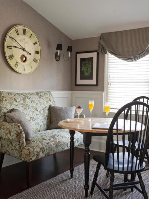 Cozy corner with a floral sofa, round table, two mimosas, and a large wall clock in a softly lit room.