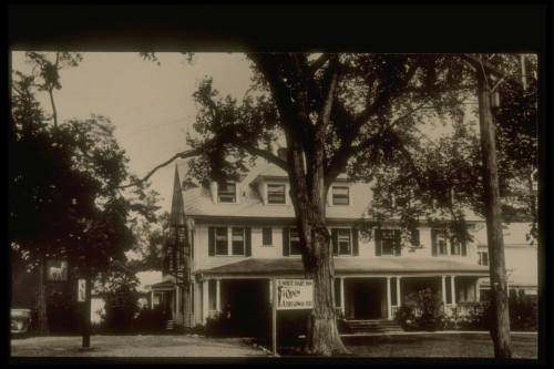 Historic black-and-white photo of a large house with a sign, surrounded by trees and a grassy area.