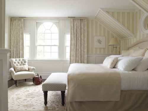 Cozy bedroom with a large bed, armchair, and elegant decor, featuring soft colors and natural light from large windows.
