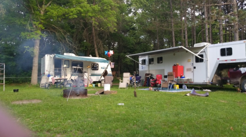 Two RVs parked in a grassy campsite with a campfire, chairs, and trees in the background. Smoke rises from the fire.