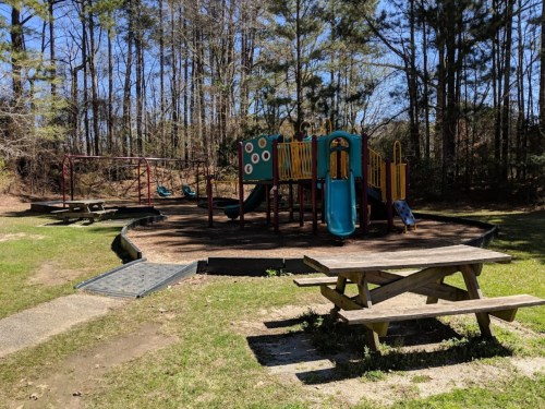 Playground with slides and swings surrounded by trees, featuring a picnic table in the foreground.
