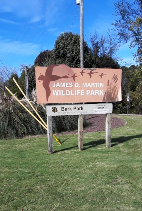 Sign for James D. Martin Wildlife Park with a lower sign for Bark Park, surrounded by greenery and power lines.
