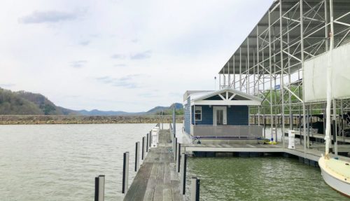 A houseboat docked at a marina, surrounded by calm water and distant hills under a cloudy sky.