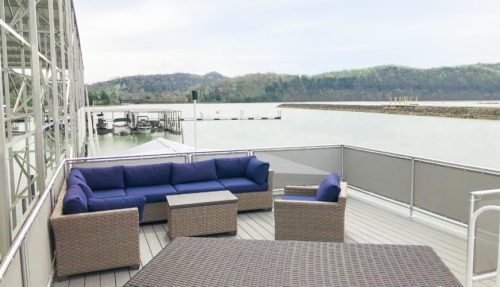A cozy outdoor seating area with a blue couch and table overlooking a calm lake and distant hills.