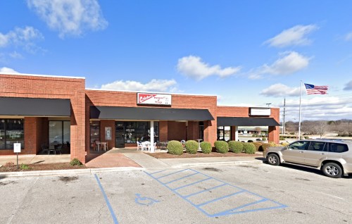 A brick storefront with awnings, featuring a sign and outdoor seating, alongside a parked SUV and an American flag.