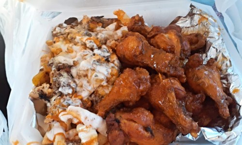 A takeout container filled with crispy chicken wings and loaded fries topped with cheese and sauce.