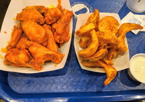 A tray with spicy chicken wings and a side of golden potato wedges, accompanied by a small cup of dipping sauce.