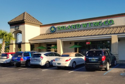 Exterior of Island Wing Co. restaurant with parked cars and palm trees under a clear blue sky.
