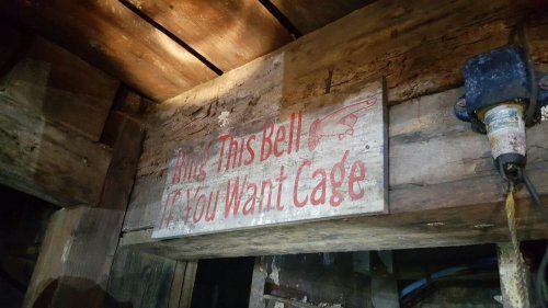 A weathered wooden sign hanging in a rustic setting, reading "Ring This Bell If You Want Cage."