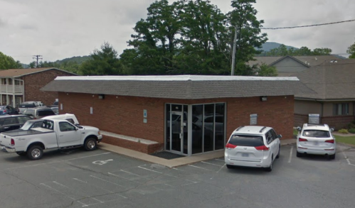 A brick building with a flat roof, surrounded by parked cars in a parking lot, set against a backdrop of trees.