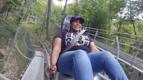 A woman smiles while riding a mountain coaster through a wooded area, wearing a black t-shirt and a cap.
