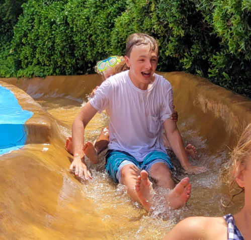 A boy joyfully slides down a water slide, splashing water around him, with greenery in the background.