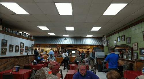 A busy diner with red-checkered tables, patrons chatting, and a kitchen visible in the background. Walls adorned with photos.
