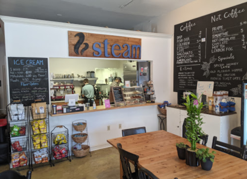 Cozy café interior with a wooden sign reading "steam," menu boards, and a display of snacks and plants.