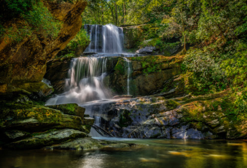 A serene waterfall cascades over rocks into a tranquil pool, surrounded by lush green trees and foliage.